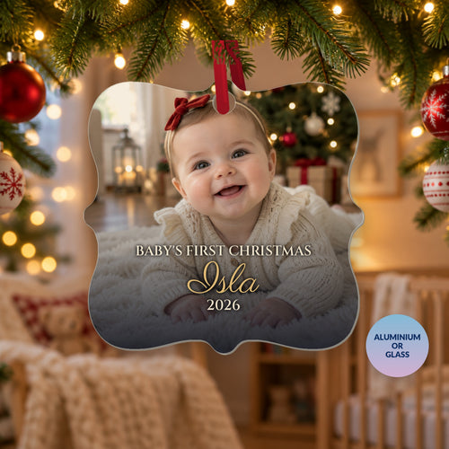 Personalised Baby's First Christmas photo ornament for Isla, 2026, with a photo of a smiling baby girl, hanging on a Christmas tree in a cosy nursery, with a badge reading Aluminium or Glass
